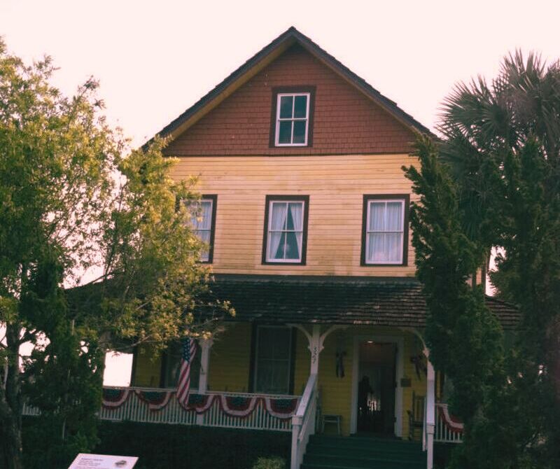 A yellow house with a flag on the porch, known as The Riddle House, steeped in a haunting legacy since 1905.