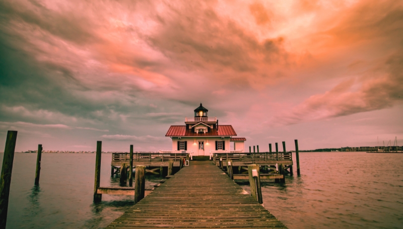 One of the most haunted places in North Carolina, the Roanoke Marshes Lighthouse is perched on a dock, surrounded by a cloudy sky, highlighting its picturesque coastal setting.