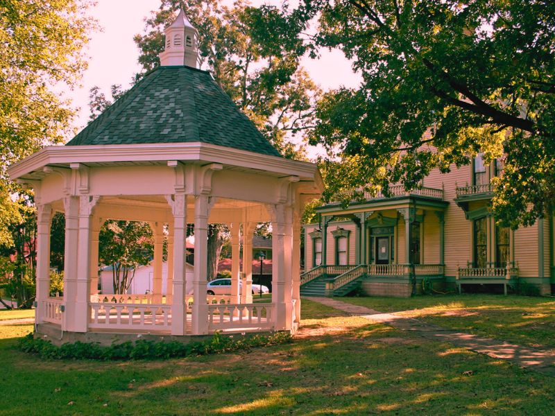 A gazebo surrounded by greenery in a park, with the haunted Clayton House visible in the background.