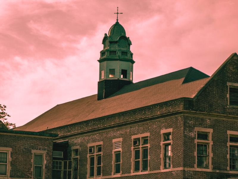 A building with a steeple, Pennhurst Asylum, set against a pink sky, famous for being one of Pennsylvania's haunted sites.