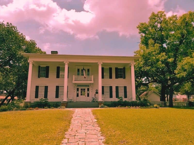The Millermore Mansion, a white historic house, set amidst a vibrant green field in Dallas, Texas.